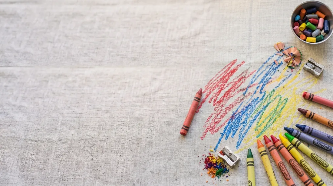 Crayon scribbles in several colours beside a box of crayons on linen