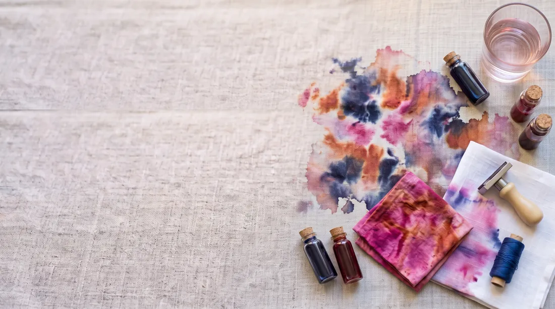 Pink and blue dye bleeding from a folded cloth onto linen, small dye bottles beside it