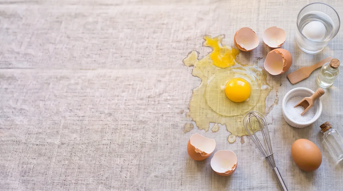 Cracked eggs with yolk and white spilled on linen, shells and a whisk nearby