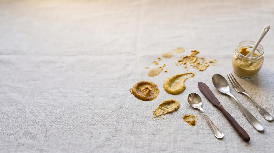 Yellow mustard smears on linen beside a mustard jar and cutlery