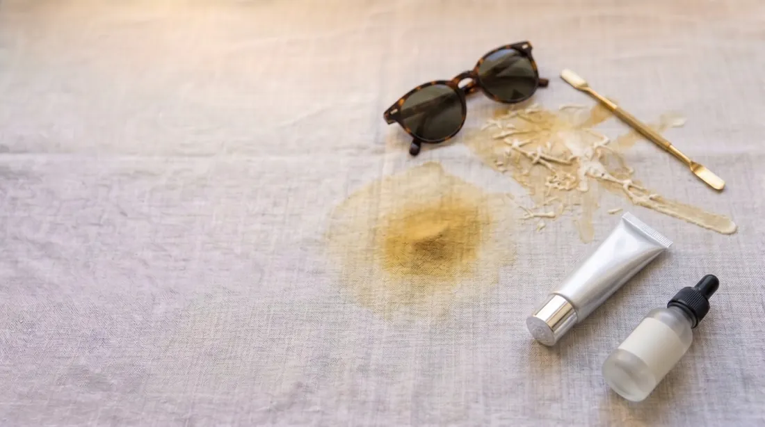 Sunscreen lotion smeared on linen beside a tube, sunglasses and a dropper bottle