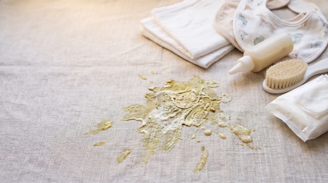 Pale vomit stain on linen beside a folded bib, bottle and cloth